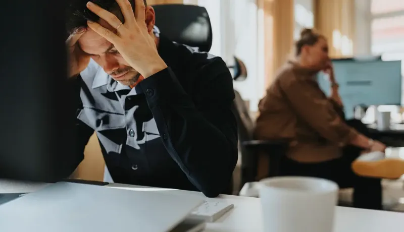 Un homme stressé, assis à un bureau, se tient la tête devant un ordinateur, dans un environnement de travail.